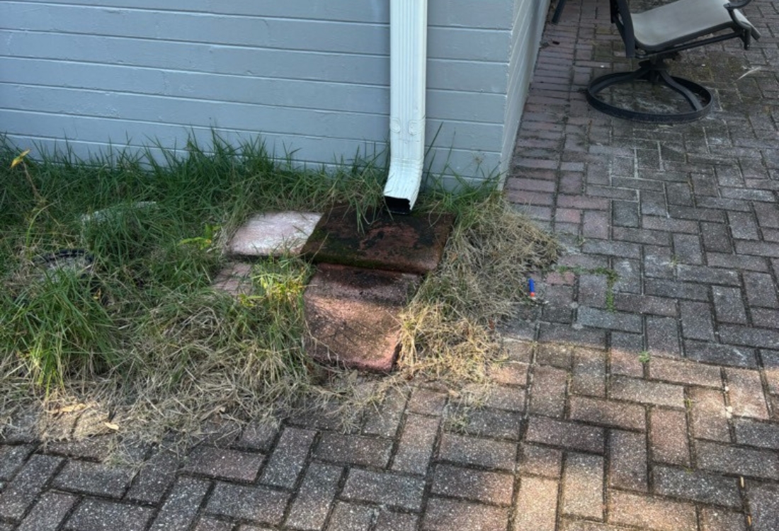 Before downspout extension, roof runoff drains at patio edge in Orange Park, FL, causing soil erosion and water damage.