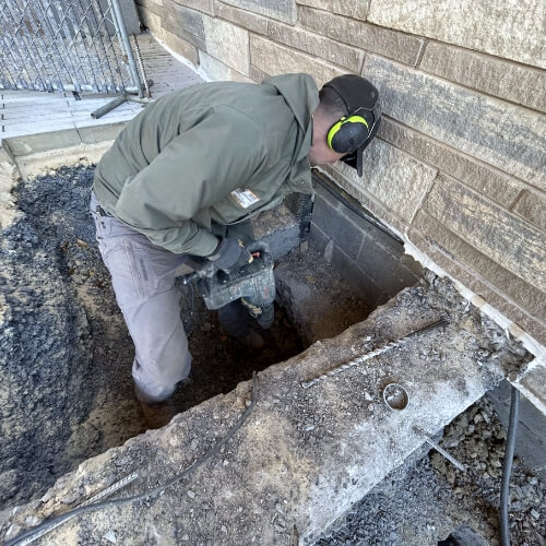 Technician excavating beside a brick wall to install push piers for foundation repair in Gainesville, Florida.