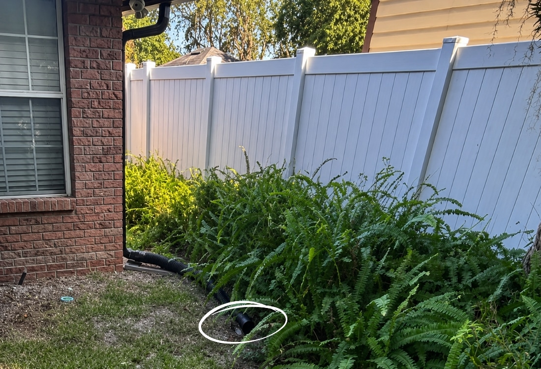 Black corrugated flex downspout discharging water near foundation base in Jacksonville, FL, causing eroded soil and water damage.