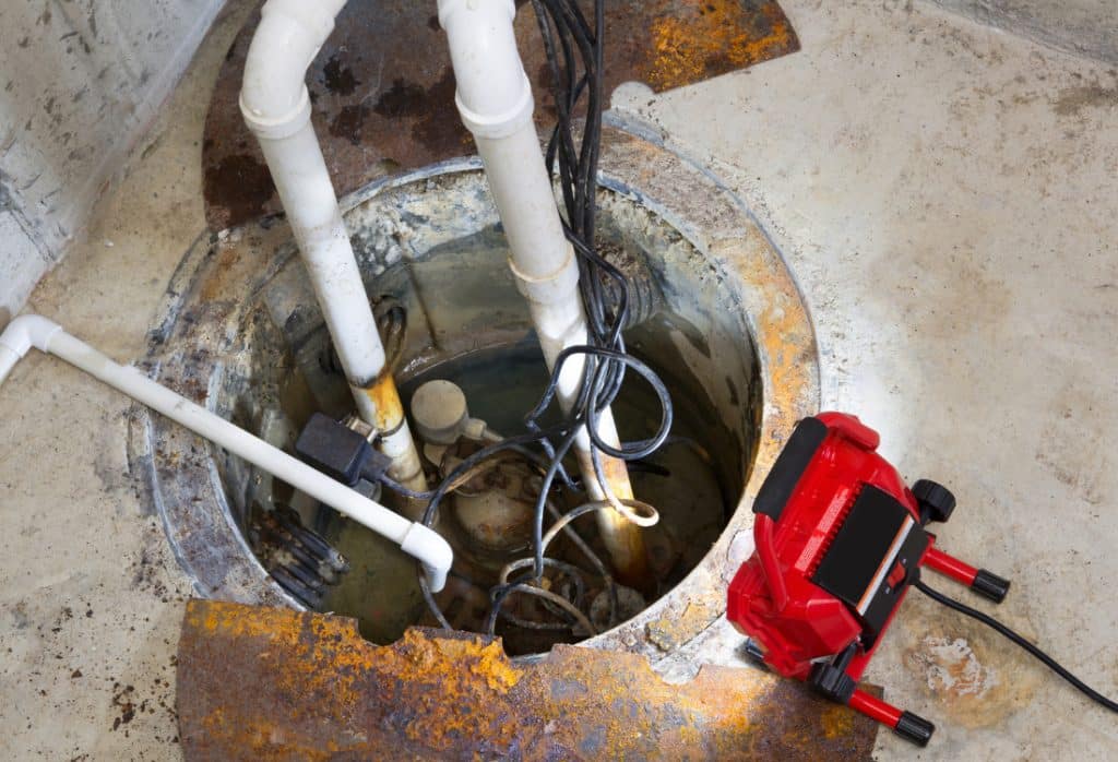 Rusted sump pump lid, corroded pipes, and tangled wiring in a basement sump pit showing signs of sump pump failure in Jacksonville, FL.
