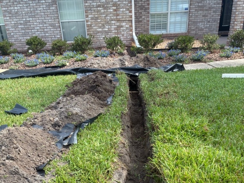 Open trench dug across the front yard during downspout drainage system installation in Jacksonville, FL.
